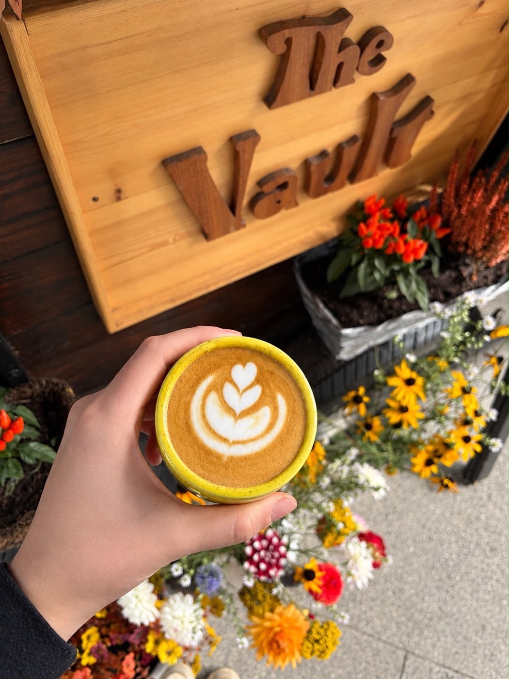 A latte with heart latte art in front of The Vault sign and flowers